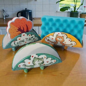 May include: Three ceramic sponge holders with cat designs. One holder is teal with the words "Love cats" and a tree-shaped sponge. Another is yellow with an orange cat design. The third is white with a green cat design.