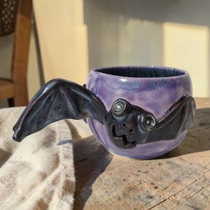 Könnte beinhalten: Ein Keramikbecher mit violetter Glasur und einer skulptierten schwarzen Fledermaus auf der Vorderseite. Die Fledermaus hat zwei hervorstehende Augen und ausgebreitete Flügel, die den Henkel bilden. Der Becher hat einen runden Körper.