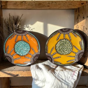 May include: Two decorative ceramic plates with a circular design. One plate has an orange background with a teal spiral center, the other has a yellow background with a green spiral center. Both plates have a brown border with a white leaf pattern.