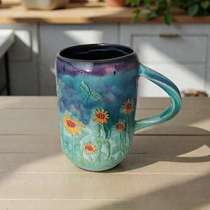 May include: A ceramic mug with a turquoise and purple glaze. The mug features embossed yellow and orange flowers with green stems and leaves. A dragonfly is also embossed on the mug. The handle and interior are a darker blue.