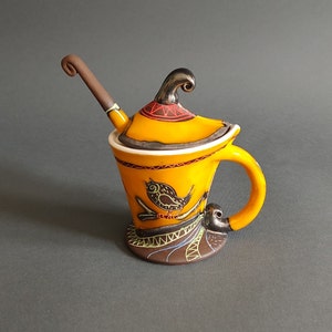 May include: A yellow ceramic creamer with a black bird design and a brown handle. The creamer has a lid with a small spout and a brown handle. The creamer is sitting on a grey surface.