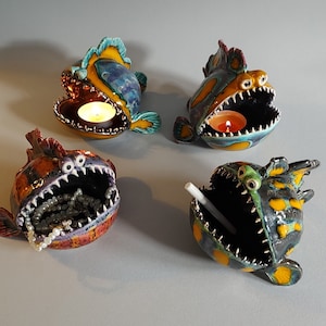 Puede incluir: Cuatro esculturas de peces de cerámica con la boca abierta, cada una con un diseño y color diferente. Los peces están decorados con pintura amarilla, naranja, azul, verde y negra. Dos de los peces tienen velas dentro de sus bocas, y uno tiene un palo blanco dentro de su boca.