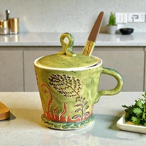 May include: A light green ceramic lidded jar with a handle and a matching spoon. The jar features a speckled finish and dark brown botanical leaf and fern illustrations around the exterior. It has a looped handle on the lid and a side handle.