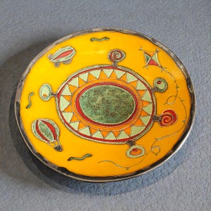 Puede incluir: Un plato decorativo redondo con un acabado de esmalte amarillo brillante y un borde plateado. El plato presenta un óvalo verde central, rodeado de triángulos naranjas y verdes. Está adornado con diseños de globos aerostáticos rojos, verdes y naranjas y remolinos caprichosos.