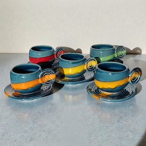 Könnte beinhalten: Ein Set aus fünf handgefertigten Keramik-Teetassen und Untertassen. Die Tassen sind türkis mit farbigen Bändern in Rot, Gelb, Orange und Grün. Jede Tasse hat einen einzigartigen Spiralgriff und steht auf einer passenden Untertasse.