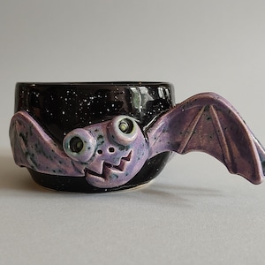 May include: A black ceramic mug with a purple bat handle. The bat has large, white eyes and a toothy grin. The mug has a speckled black glaze.