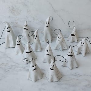 May include: A collection of thirteen white ceramic ghost ornaments, each featuring a unique hand-painted facial expression and a black string loop for hanging. The ghosts vary in size and shape, with some having smooth surfaces and others featuring dark speckles.