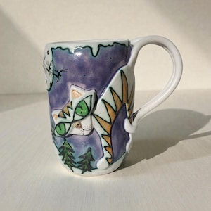 Puede incluir: Una taza de cerámica con un asa blanca y un diseño caprichoso de gato. La taza presenta un fondo morado con un gato blanco, ojos verdes y una escena de bosque. La taza está pintada a mano con un acabado brillante.