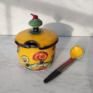 Könnte beinhalten: Eine gelbe Zuckerdose aus Keramik mit passendem Löffel. Die Dose hat einen Deckel mit einer grünen und roten Spitze. Dose und Löffel sind mit farbenfrohen, skurrilen Designs verziert. Der Löffel ist 15 cm lang.