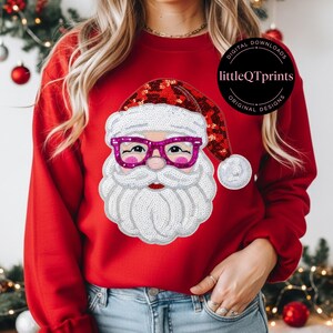 Op de afbeelding: Een rode sweatshirt met een kerstmanontwerp met een pailletten hoed en baard, en een roze bril. Het gezicht van de kerstman is gemaakt van witte pailletten. Op de sweatshirt staat de tekst "littleQTprints".