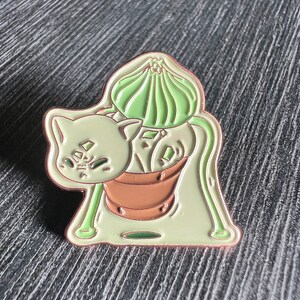 May include: A green and brown enamel pin featuring a cartoon cat with a plant growing out of its head. The plant is in a brown pot and has a green flower on top. The cat is looking to the left and has a surprised expression on its face.