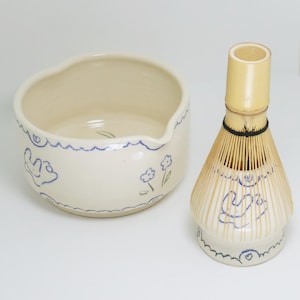 Peut inclure: Un ensemble de bol à matcha et de fouet en bambou de couleur crème. Le bol a un bord ondulé et est décoré de dessins au trait bleus de fleurs et de formes abstraites. Le fouet a un manche et des brins en bambou.
