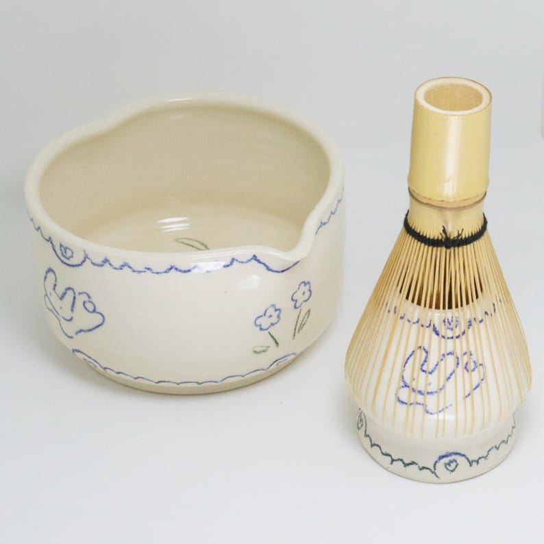 Puede incluir: Un juego de cuenco de matcha y batidor de bamb&uacute; de color crema. El cuenco tiene un borde ondulado y est&aacute; decorado con dibujos lineales azules de flores y formas abstractas. El batidor tiene un mango y p&uacute;as de bamb&uacute;.
