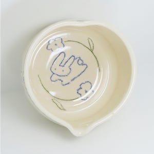 Puede incluir: Un peque&ntilde;o cuenco de cer&aacute;mica blanquecina, de forma redondeada y con un pico vertedor. El interior presenta un boceto azul de un conejo rodeado de flores y hojas verdes. Ideal para servir salsas o aperitivos.
