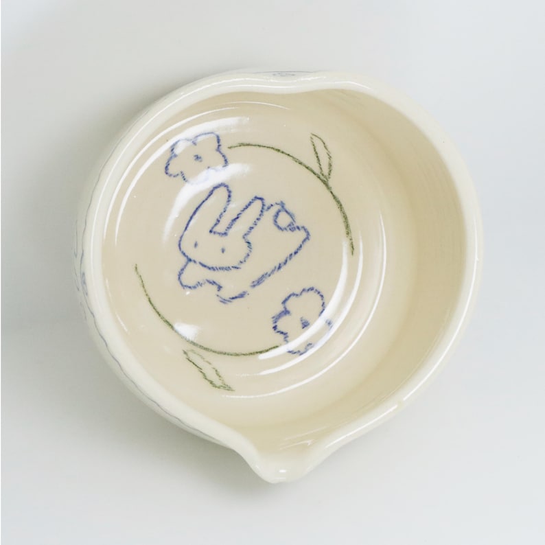 Puede incluir: Un peque&ntilde;o cuenco de cer&aacute;mica blanquecina, de forma redondeada y con un pico vertedor. El interior presenta un boceto azul de un conejo rodeado de flores y hojas verdes. Ideal para servir salsas o aperitivos.