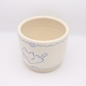Puede incluir: Una taza de cer&aacute;mica color crema con un dibujo en azul de un animal y corazones. El borde y la base de la taza tienen un dise&ntilde;o festoneado. El interior de la taza es de color beige claro.