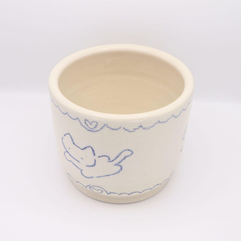 Puede incluir: Una taza de cer&aacute;mica color crema con un dibujo en azul de un animal y corazones. El borde y la base de la taza tienen un dise&ntilde;o festoneado. El interior de la taza es de color beige claro.