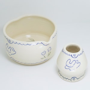 Puede incluir: Dos piezas de cer&aacute;mica blanquecina con dise&ntilde;os azules dibujados a mano. Un cuenco con borde ondulado y un peque&ntilde;o jarr&oacute;n con forma de hongo. Ambos tienen patrones florales y abstractos azules.