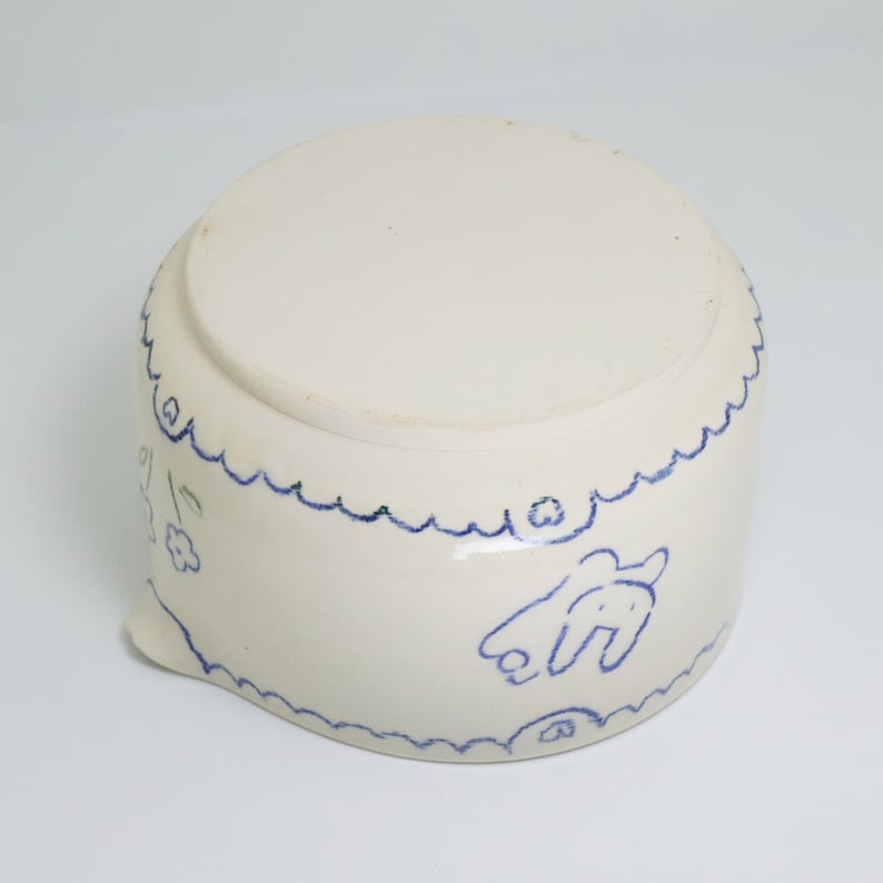 Puede incluir: Un cuenco de cer&aacute;mica blanca con un borde azul festoneado e ilustraciones azules de un conejo y flores. El cuenco tiene un peque&ntilde;o pico y probablemente est&aacute; hecho a mano. Es un utensilio de cocina &uacute;nico.