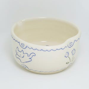 Puede incluir: Cuenco de cer&aacute;mica color crema con un borde ondulado azul y una ilustraci&oacute;n de gato azul. El cuenco tambi&eacute;n presenta un peque&ntilde;o dise&ntilde;o floral. El cuenco tiene un peque&ntilde;o pico en el lateral.