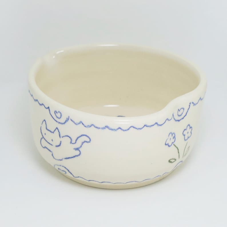 Puede incluir: Cuenco de cer&aacute;mica color crema con un borde ondulado azul y una ilustraci&oacute;n de gato azul. El cuenco tambi&eacute;n presenta un peque&ntilde;o dise&ntilde;o floral. El cuenco tiene un peque&ntilde;o pico en el lateral.