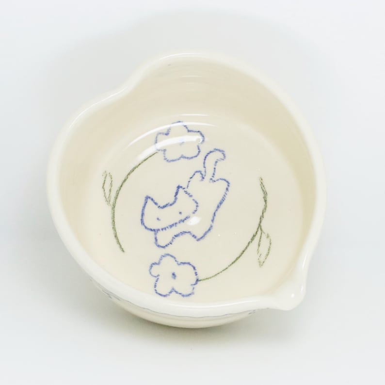 Puede incluir: Un peque&ntilde;o cuenco de cer&aacute;mica blanquecina con borde en forma de coraz&oacute;n. El interior presenta un dise&ntilde;o caprichoso de gato y flores azules, con tallos y hojas verdes. El cuenco tiene un peque&ntilde;o pico en un lado.