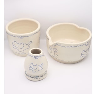 Puede incluir: Un conjunto de tres piezas de cer&aacute;mica blanquecina con dise&ntilde;os de gatos y corazones azules. El conjunto incluye un jarr&oacute;n peque&ntilde;o, un cuenco redondo y un cuenco en forma de coraz&oacute;n. Los bordes de cada pieza tienen una l&iacute;nea azul ondulada.