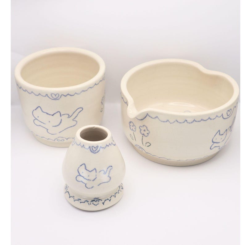 Puede incluir: Un conjunto de tres piezas de cer&aacute;mica blanquecina con dise&ntilde;os de gatos y corazones azules. El conjunto incluye un jarr&oacute;n peque&ntilde;o, un cuenco redondo y un cuenco en forma de coraz&oacute;n. Los bordes de cada pieza tienen una l&iacute;nea azul ondulada.