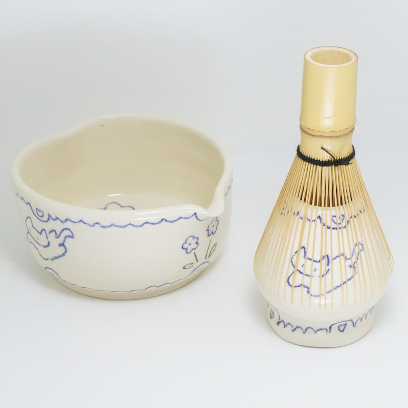 Puede incluir: Un juego de cuenco de matcha y batidor de bamb&uacute;. El cuenco blanquecino tiene un borde ondulado y est&aacute; decorado con dibujos lineales azules de flores y formas abstractas. El batidor es de color beige claro con una banda negra y dibujos lineales azules.