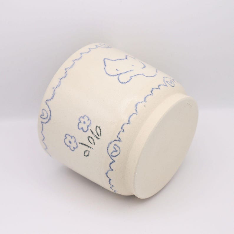 Puede incluir: Una maceta de cer&aacute;mica color crema con dise&ntilde;os florales y de oso azules dibujados a mano. La maceta tiene un borde azul festoneado y es adecuada para plantas peque&ntilde;as u objetos decorativos. La maceta mide aproximadamente 10 cm de alto.