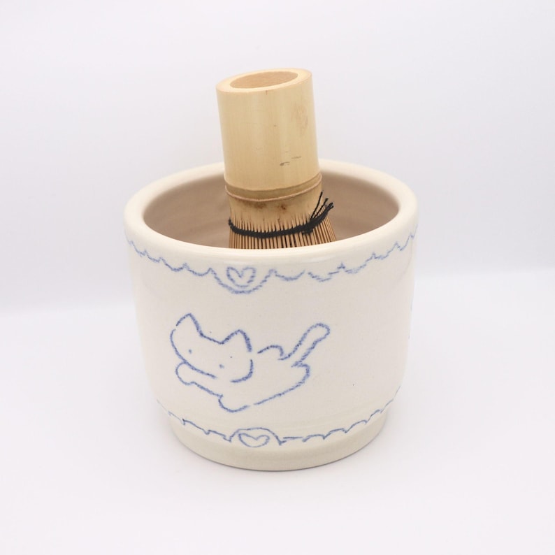 Puede incluir: Un cuenco de cer&aacute;mica blanca para matcha con una ilustraci&oacute;n de gato azul y detalles de coraz&oacute;n, que contiene un batidor de matcha de bamb&uacute;. El batidor es de bamb&uacute; natural, para preparar t&eacute; matcha.