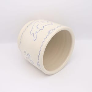 Puede incluir: Una taza de cer&aacute;mica color crema con un dise&ntilde;o de contorno azul. El dise&ntilde;o incluye un conejito y motivos florales. La taza tiene forma cil&iacute;ndrica y un acabado liso y brillante. El interior es de color beige claro.