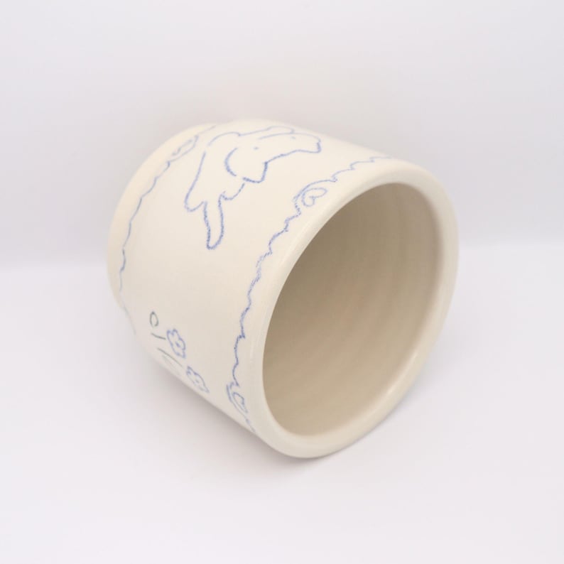 Puede incluir: Una taza de cer&aacute;mica color crema con un dise&ntilde;o de contorno azul. El dise&ntilde;o incluye un conejito y motivos florales. La taza tiene forma cil&iacute;ndrica y un acabado liso y brillante. El interior es de color beige claro.
