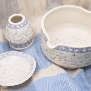 May include: A ceramic set consisting of a small rounded vase, a shallow dish, and a wide bowl with a pouring spout. Each piece is off-white with a blue hand-drawn pattern featuring stylized cats, small stars, and geometric border designs.
