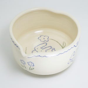 Puede incluir: Cuenco de cer&aacute;mica blanquecina con pico, decorado con dibujos lineales azules de flores y un conejito. El borde tiene una l&iacute;nea azul ondulada. El cuenco tiene una est&eacute;tica caprichosa y hecha a mano.