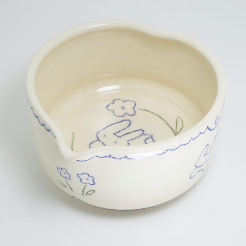 Puede incluir: Cuenco de cer&aacute;mica blanquecina con pico, decorado con dibujos lineales azules de flores y un conejito. El borde tiene una l&iacute;nea azul ondulada. El cuenco tiene una est&eacute;tica caprichosa y hecha a mano.