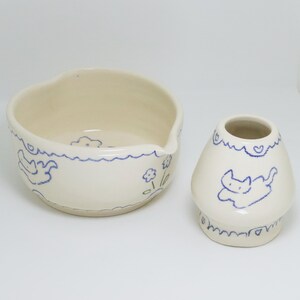 Puede incluir: Dos piezas de cer&aacute;mica blanquecina con dibujos lineales azules. Un cuenco con borde ondulado y pico, y un peque&ntilde;o jarr&oacute;n con forma de hongo. Ambos presentan dise&ntilde;os de gatos y flores.