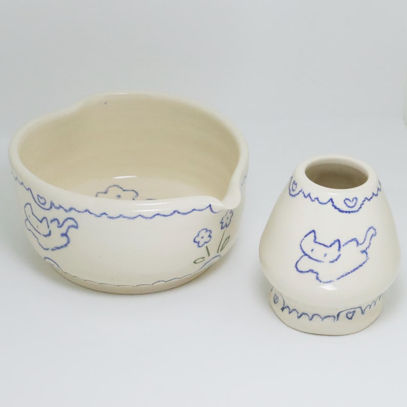 Puede incluir: Dos piezas de cer&aacute;mica blanquecina con dibujos lineales azules. Un cuenco con borde ondulado y pico, y un peque&ntilde;o jarr&oacute;n con forma de hongo. Ambos presentan dise&ntilde;os de gatos y flores.