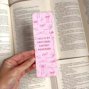 May include: Pink bookmark with a white border and a pink and white bow pattern. The bookmark has the text "This is my emotional support bookmark" in red and white.