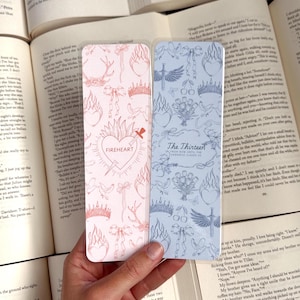May include: Two bookmark designs with a pink and blue background. The pink bookmark features a heart with a sword through it and the text "FIREHEART". The blue bookmark features a bouquet of flowers and the text "The Thirteen From Now Until The Darkness Claims Us".