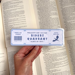May include: A blue and white bookmark with the text "Basgiath War College Riders Quadrant Class of 2013" and "admit one student" with a barcode. The bookmark also has a dragon graphic and the text "status: first year".