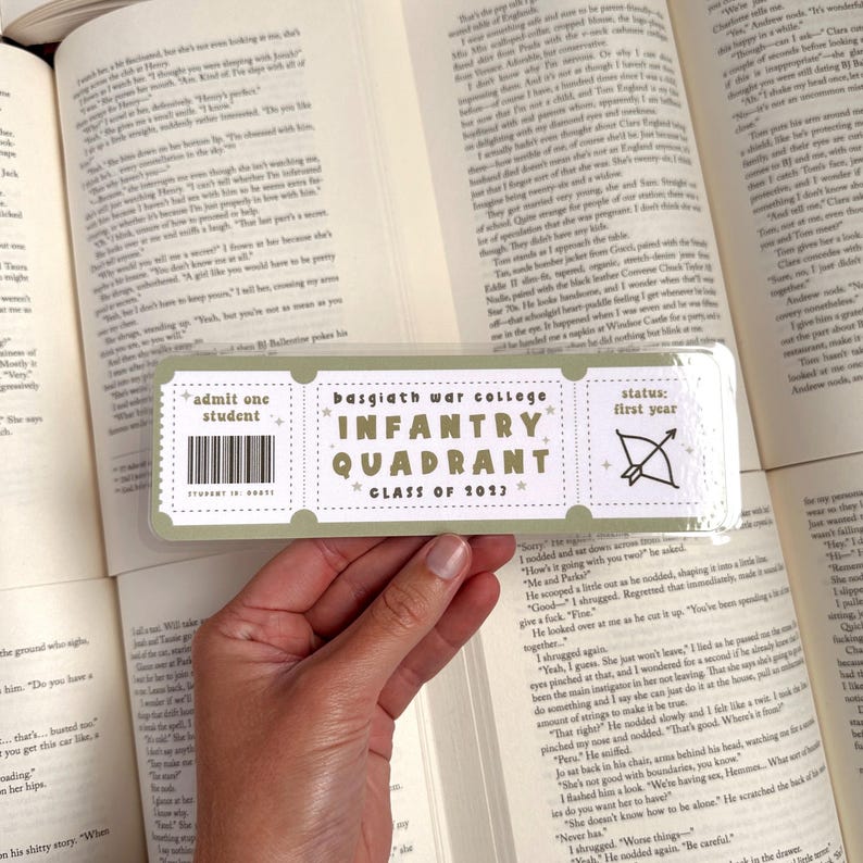 May include: A green and white bookmark with a ticket design. The bookmark says "Basgiath War College Infantry Quadrant Class of 1013" and "Status: First Year". The bookmark has a white arrow and bow on it.