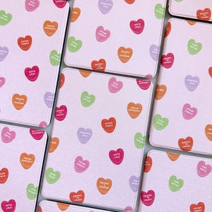 Puede incluir: Un juego de ocho posavasos rosas con fondo blanco y un patr&oacute;n repetido de caramelos en forma de coraz&oacute;n de colores con diferentes lemas. Los lemas incluyen "Soul Mates", "Second Chance", "Found Family", "Grumpy & Jealous", "Insta Love", "Forced Proximity", "Slow Burn", "Free Dating", "Enemies to Lovers", "Pre-Pregnant", "Friends to Lovers", "Karma to Lovers", "Loveas", "Fare Rating", "What", "100% Mates".