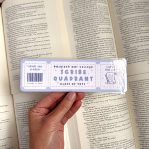May include: A light blue bookmark with a ticket design. It says "admit one student" on the left, "basgiath war college *SCRIBE* QUADRANT CLASS OF 2023" in the middle, and "stat's first year" on the right.