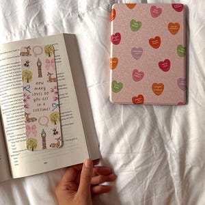 Puede incluir: Un marcap&aacute;ginas rosa con fondo blanco e ilustraciones coloridas de un ciervo, una abeja, una torre del reloj y un &aacute;rbol. El marcap&aacute;ginas tiene el texto "&iquest;Cu&aacute;ntos amores se consiguen en una vida?" impreso en &eacute;l.