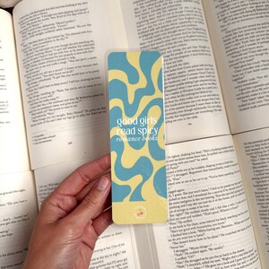 May include: A yellow and blue bookmark with a wavy abstract design. The bookmark has the text "good girls read spicy romance books."