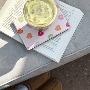 Puede incluir: Una bebida de matcha verde en un vaso de pl&aacute;stico transparente con tapa, sentada encima de un libro rosa y blanco con un patr&oacute;n de corazones. El libro est&aacute; abierto y el texto "The Twisted Crowns" es visible en la p&aacute;gina. El libro est&aacute; sentado en un sof&aacute; gris.