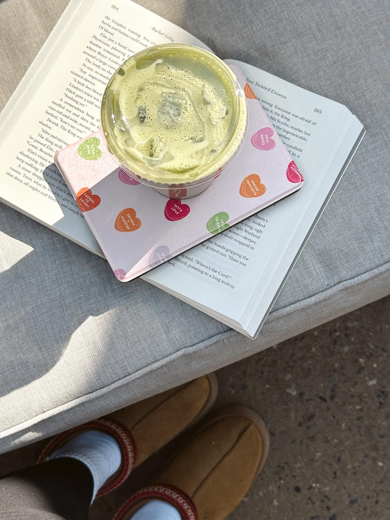 Puede incluir: Una bebida de matcha verde en un vaso de pl&aacute;stico transparente con tapa, sentada encima de un libro rosa y blanco con un patr&oacute;n de corazones. El libro est&aacute; abierto y el texto "The Twisted Crowns" es visible en la p&aacute;gina. El libro est&aacute; sentado en un sof&aacute; gris.