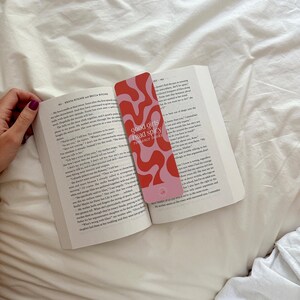 May include: A pink and red bookmark with the text "good girls read spicy romance books" and abstract shapes. The bookmark is laying on top of an open book.