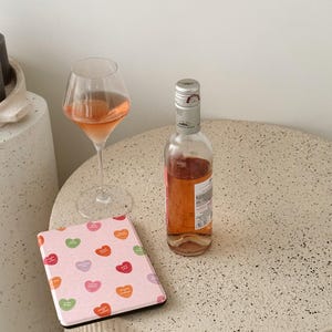 Puede incluir: Un lector electr&oacute;nico rosa con un patr&oacute;n de corazones coloridos, una copa de vino rosado y una botella de vino rosado sobre una superficie beige moteada.